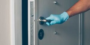 Hand wearing a blue glove pushing down the metallic handle of an open modern door with a multi-point locking mechanism.